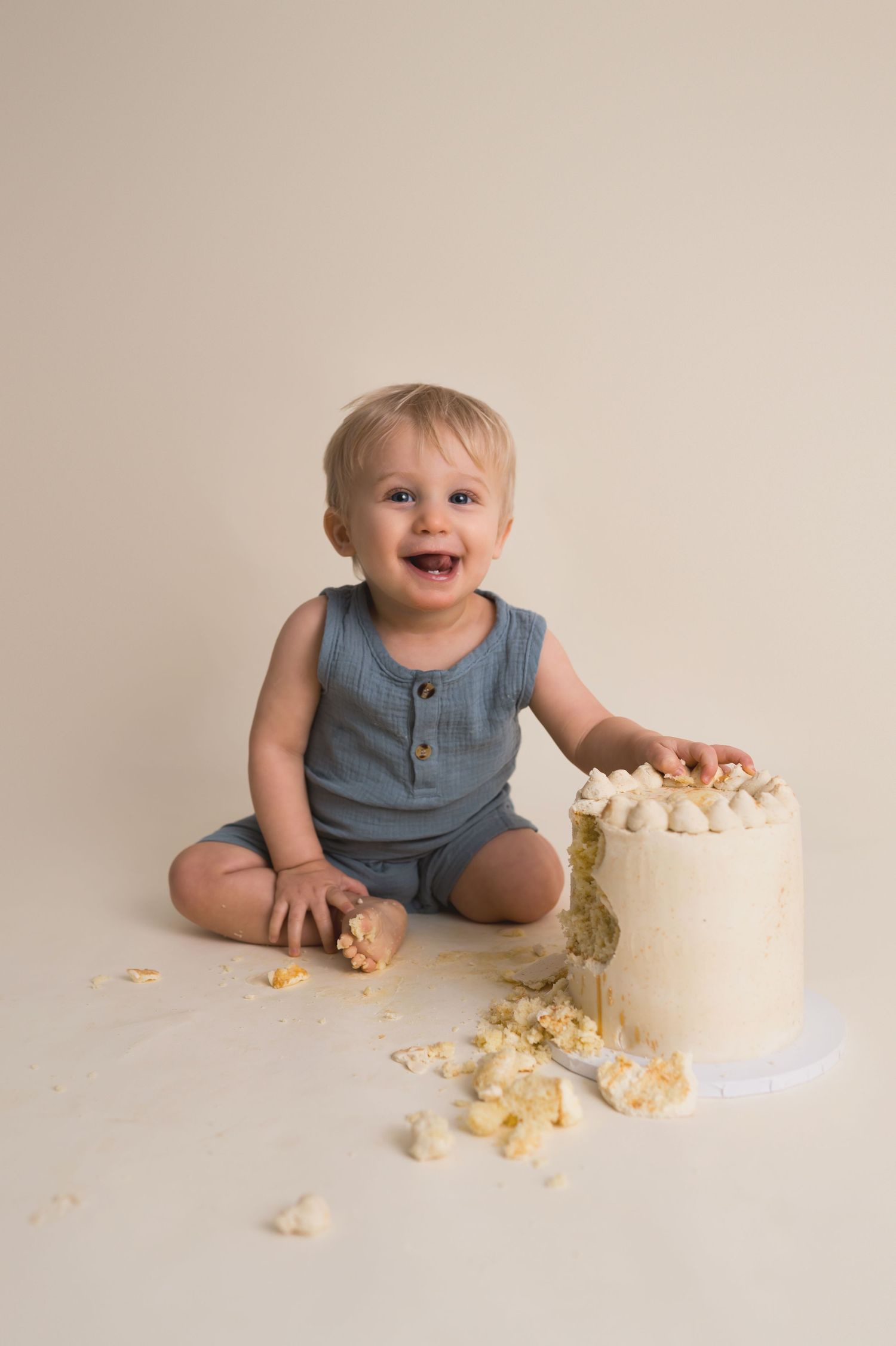 Happy baby in blue romper sits on floor next to white smash cake with crumbs scattered around.