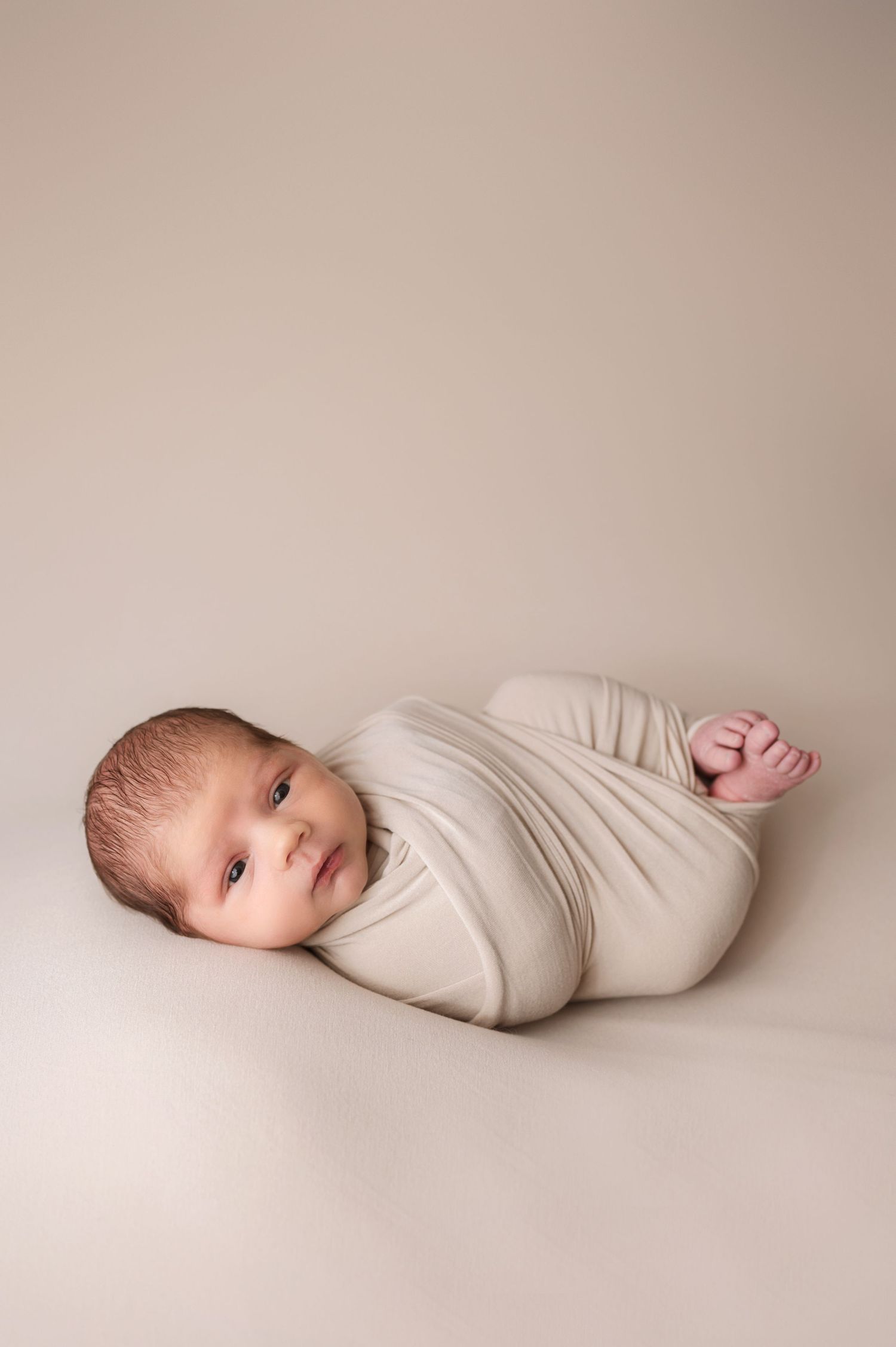 Newborn baby swaddled in cream fabric lying on beige background during professional photography session.